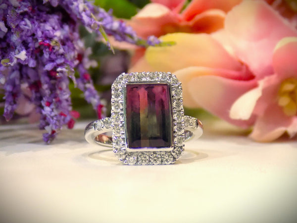 This is a women's ring with a natural 2.66ct bi-color tourmaline. It's colors are a deep rich blueish green on one end that turns to a vivid pink on the other side. The tourmaline is surrounded by .66ct in natural round accent diamonds in 14K white gold on a white surface with a multi color floral background.