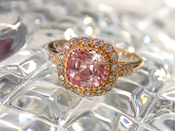 This is a women's ring with 1.94ct natural cushion cut morganite in the center surrounded by .30ct in natural round diamond accents in 14K rose gold sitting on a textured glass surface.