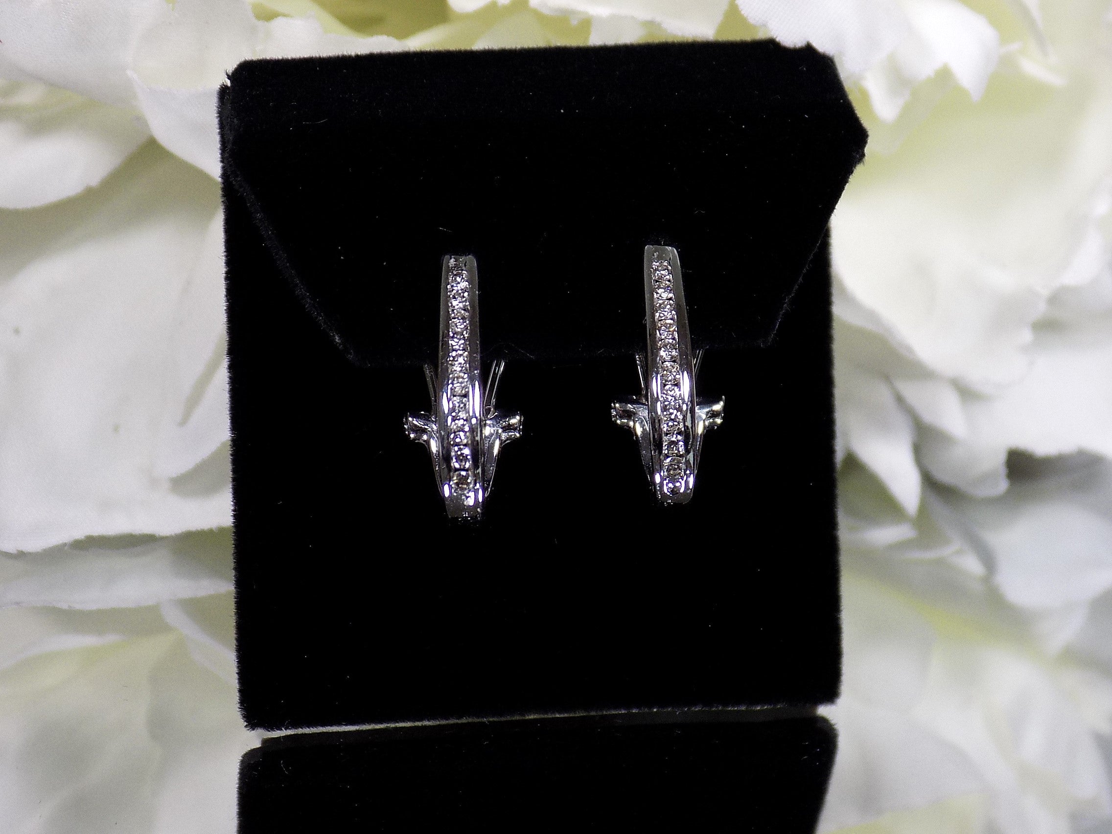 "J" hoop .25ct natural diamond earrings channel set in 10K white gold on a black pad with a white floral background.