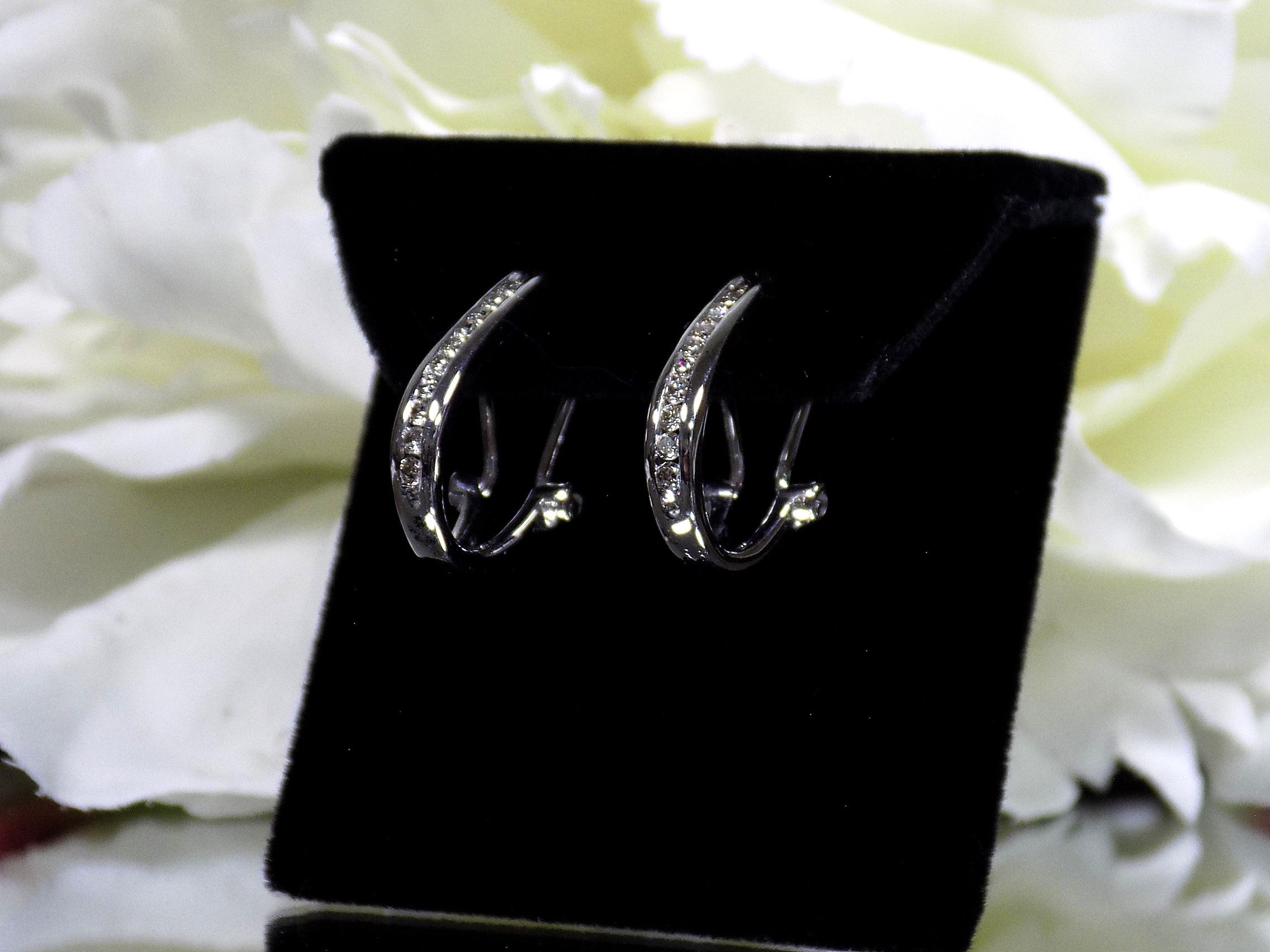 "J" hoop .25ct natural diamond earrings channel set in 10K white gold on a black pad with a white floral background. Side view.