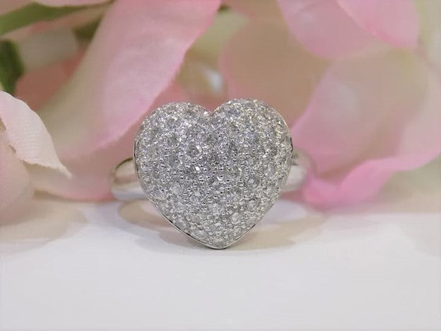 This is a 1.00ct heart shaped pave set natural diamond ring in 18K white gold on a mirrored surface with a pink floral background.