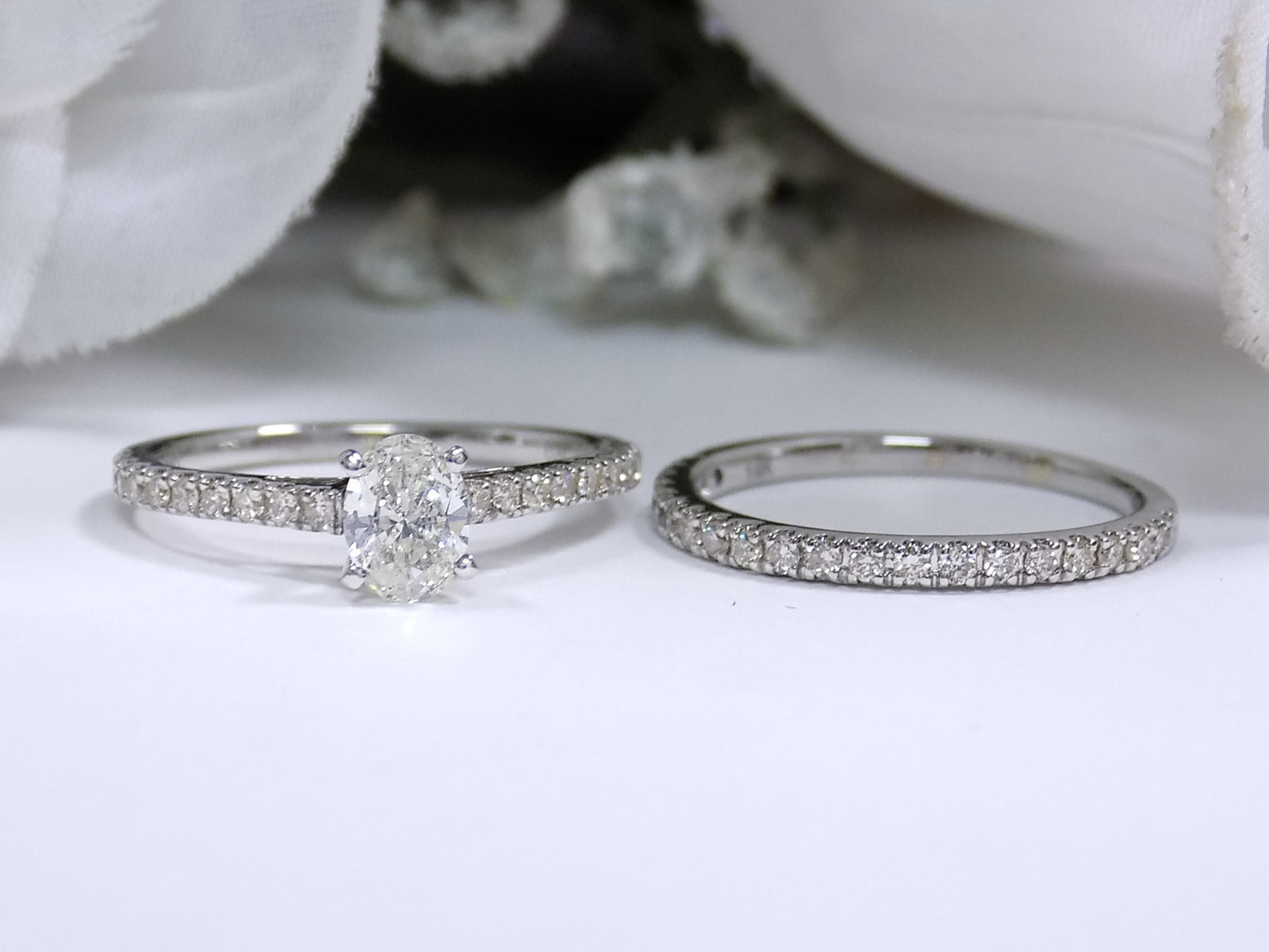 This shows both pieces of an oval shape diamond wedding set - the engagement ring laying beside its matching band with a white gold.