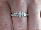 This is a marquise shape diamond ring with round diamond accents in white gold shown on a ladies' finger.