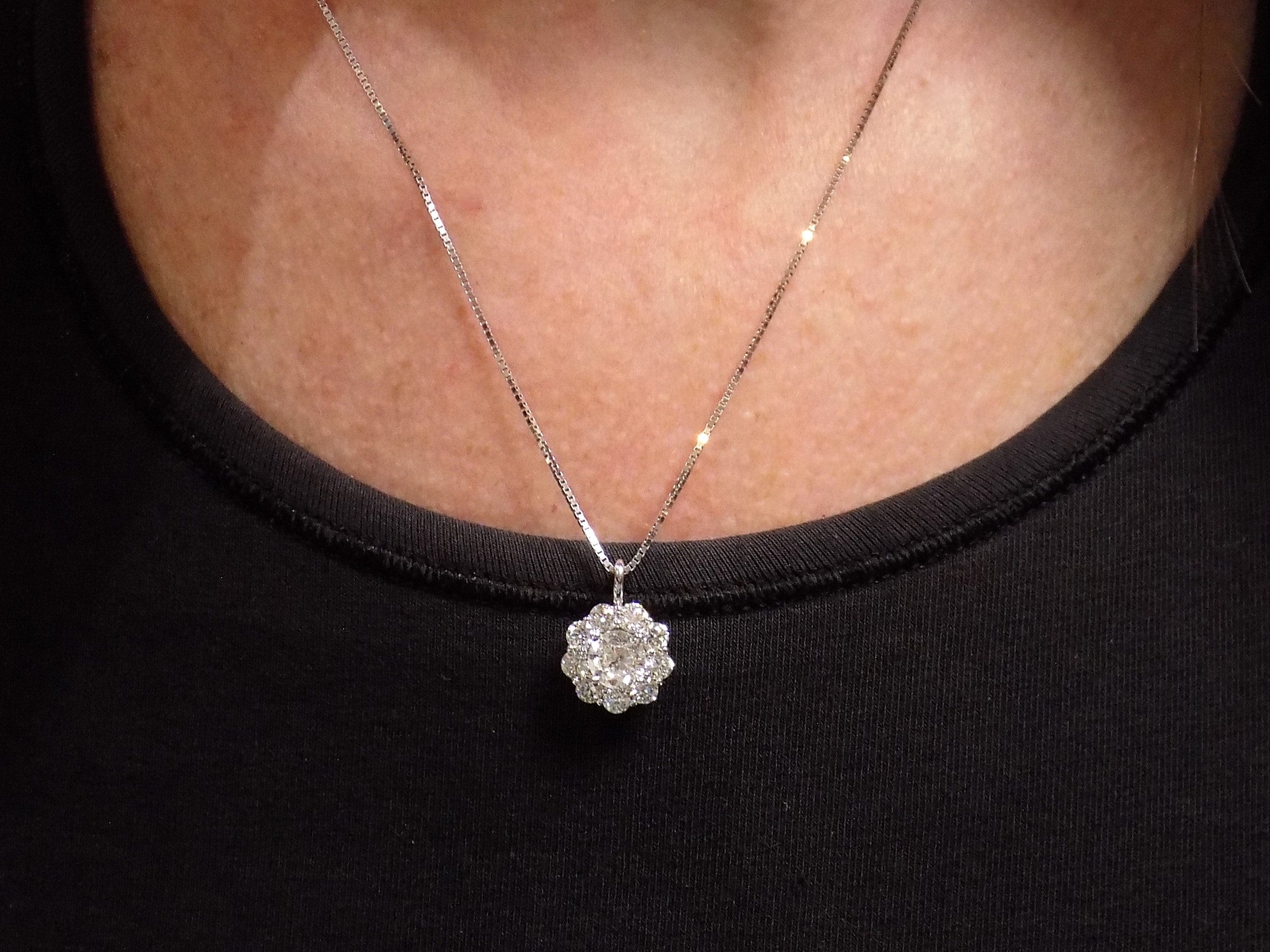 This is a 2.00ct natural round halo style diamond necklace with a 1.00ct center round brilliant diamond and 1.00ct round brilliant diamonds in the halo in 14K white gold on a ladies neck with a black top.