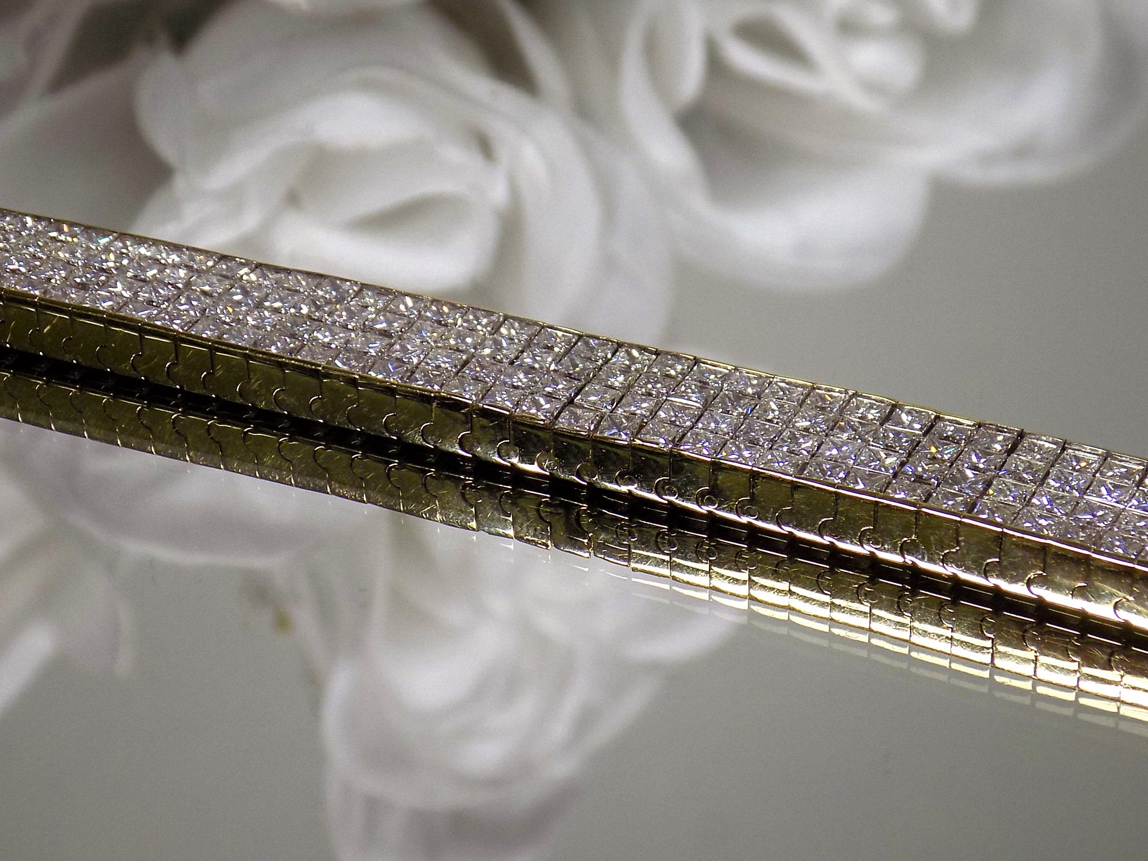 This is a 22.00ct natural princess cut diamond bracelet with 76.4 grams of 18K yellow gold on a mirrored surface with a white floral background.
