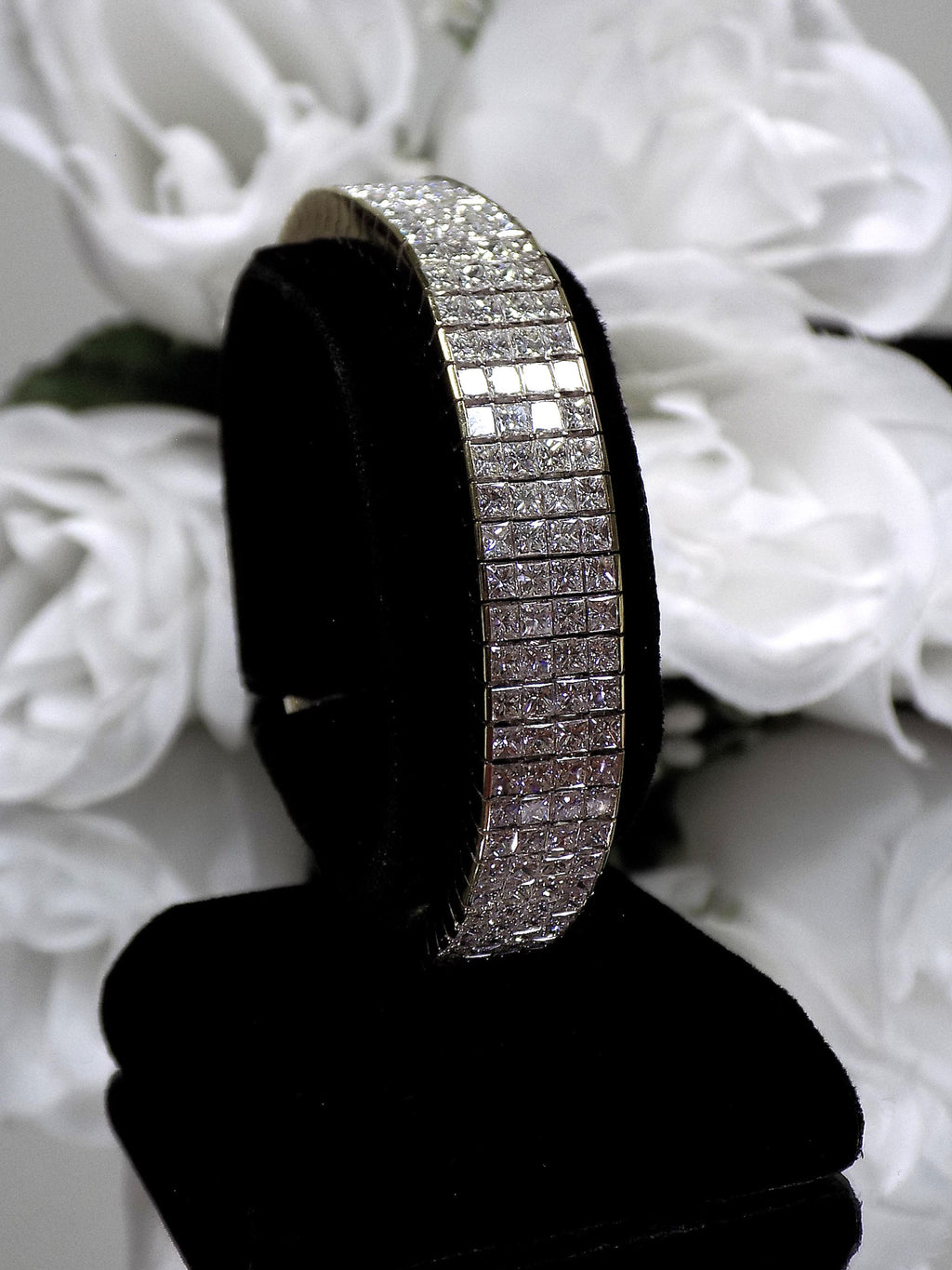 This is a 22.00ct natural princess cut diamond bracelet with 76.4 grams of 18K yellow gold on black stand with a white floral background.