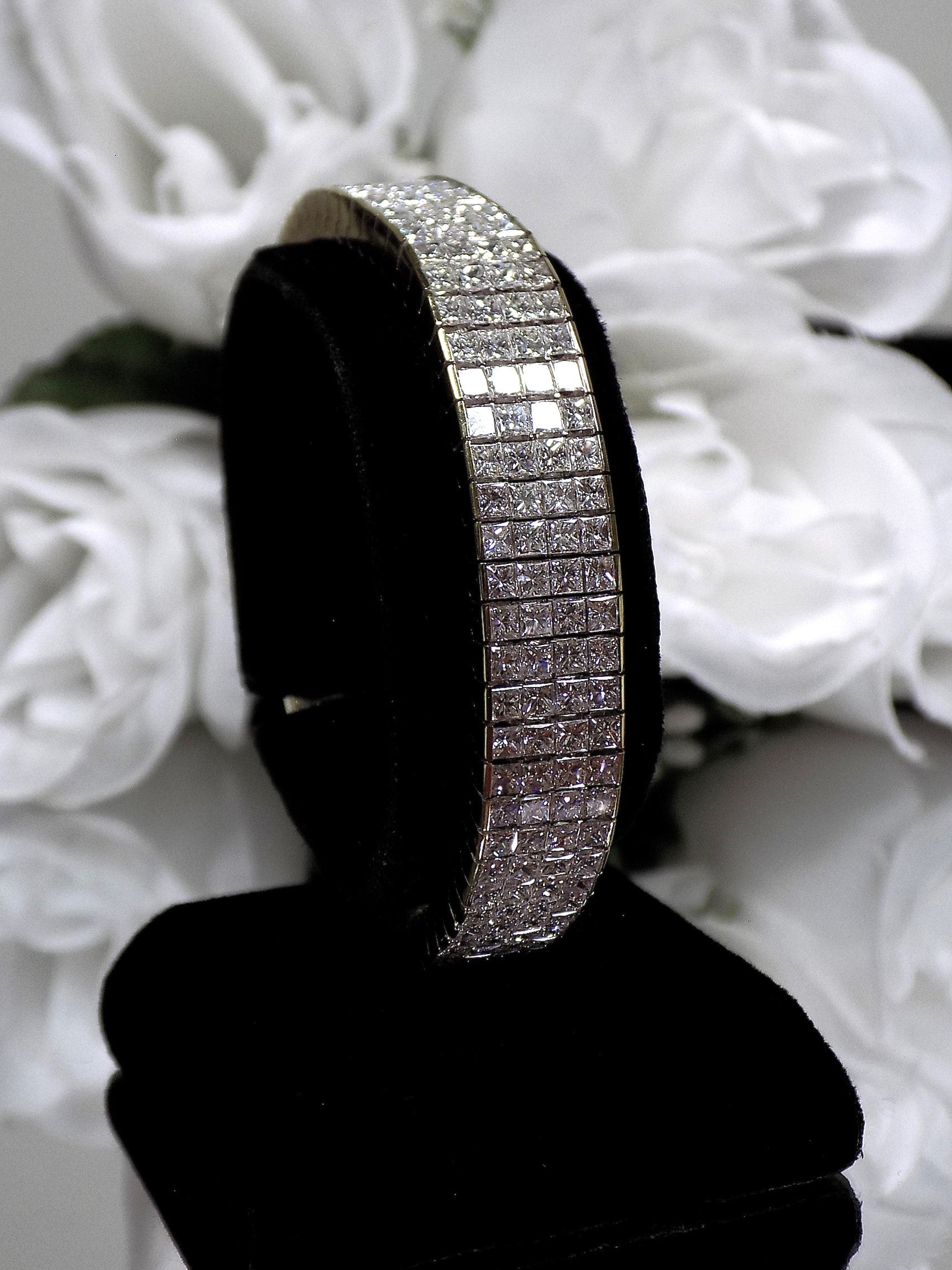 This is a 22.00ct natural princess cut diamond bracelet with 76.4 grams of 18K yellow gold on black stand with a white floral background.