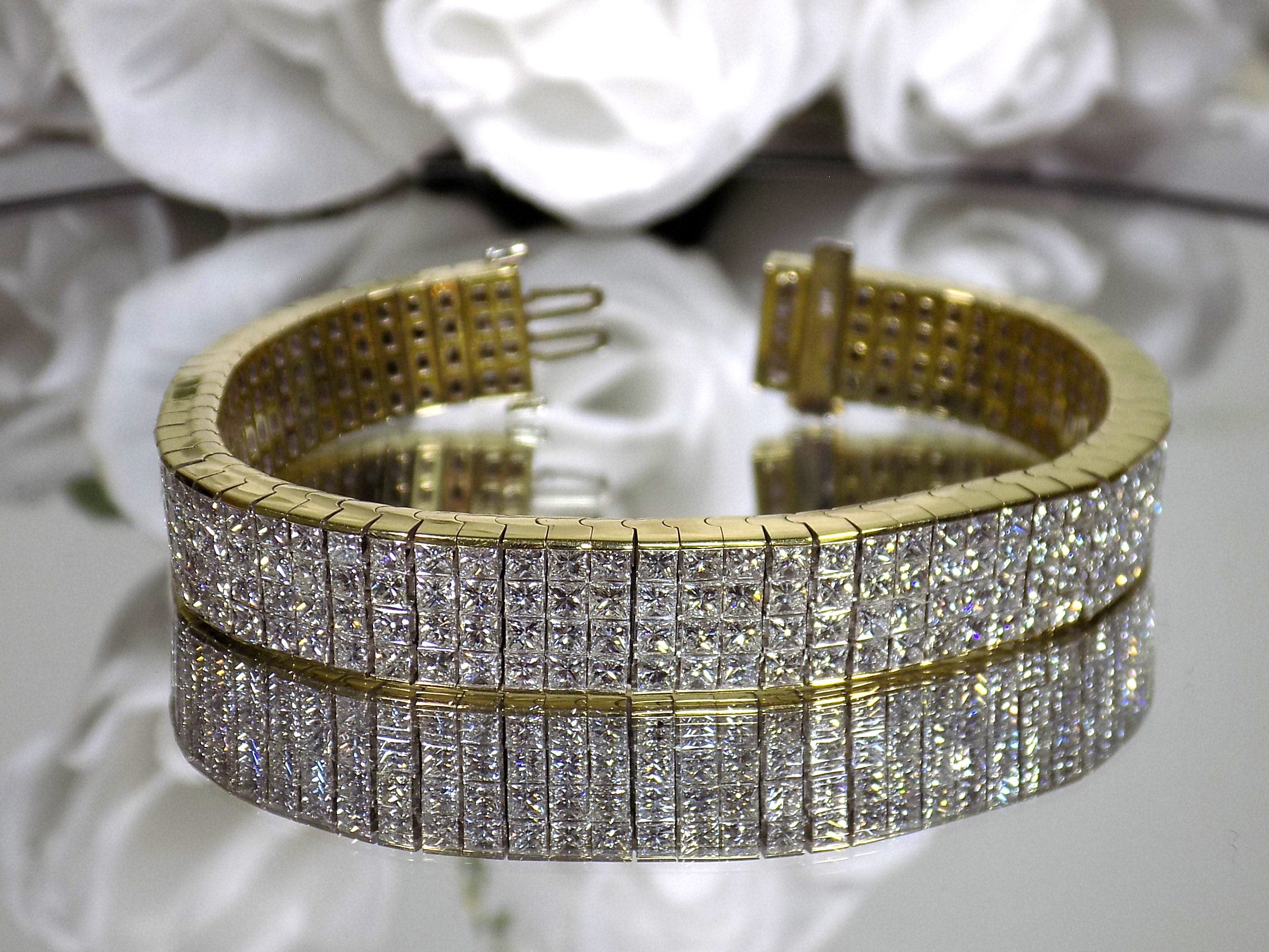 This is a 22.00ct natural princess cut diamond bracelet with 76.4 grams of 18K yellow gold on a mirrored surface with a white floral background.