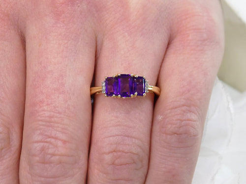 This is a 1.25ct total weight 3-stone natural amethyst and round diamond  ring in 10K yellow gold on a woman's finger.
