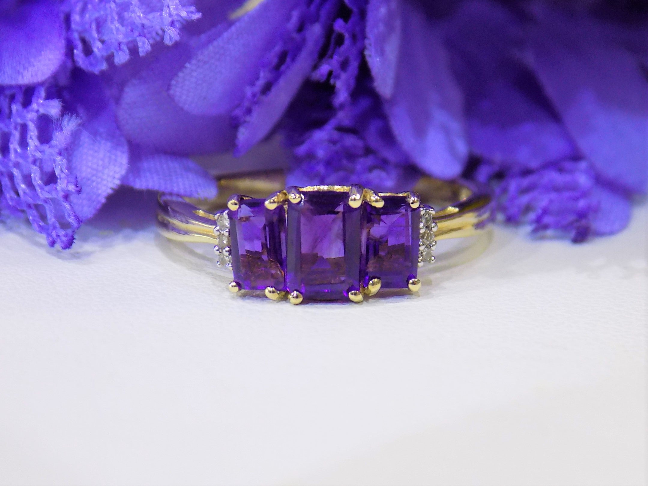 This is a 1.25ct total weight 3-stone natural amethyst and round diamond  ring in 10K yellow gold with a white and purple floral background.