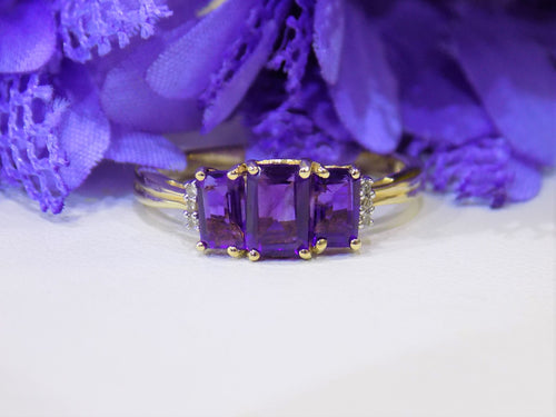 This is a 1.25ct total weight 3-stone natural amethyst and round diamond  ring in 10K yellow gold with a white and purple floral background.