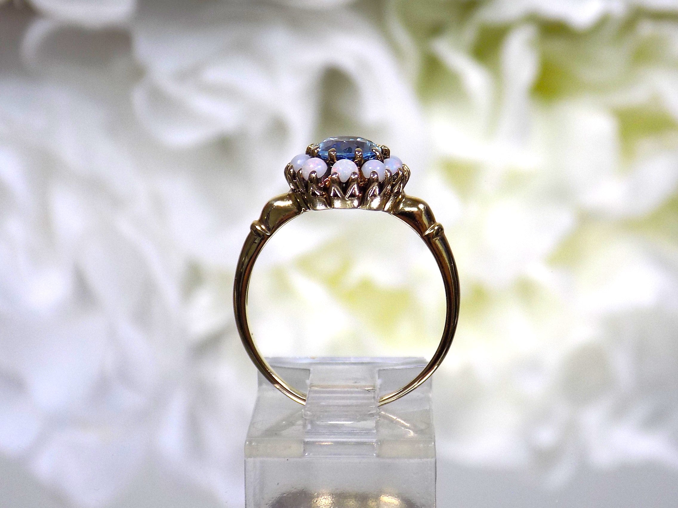This a side view of a ring with an oval blue topaz in the center and surrounded by natural opals in yellow gold from our estate collection with a flowery background.