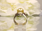 This is a women's vintage ring with center 9mm golden akoya cultured pearl and 2 .25ct bezel set natural rubies in 18K yellow gold on a mirrored surface with and white floral background from a side view.