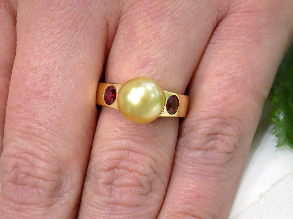 This is a women's vintage ring with center 9mm golden akoya cultured pearl and 2 .25ct bezel set natural rubies in 18K yellow gold on a women's finger.
