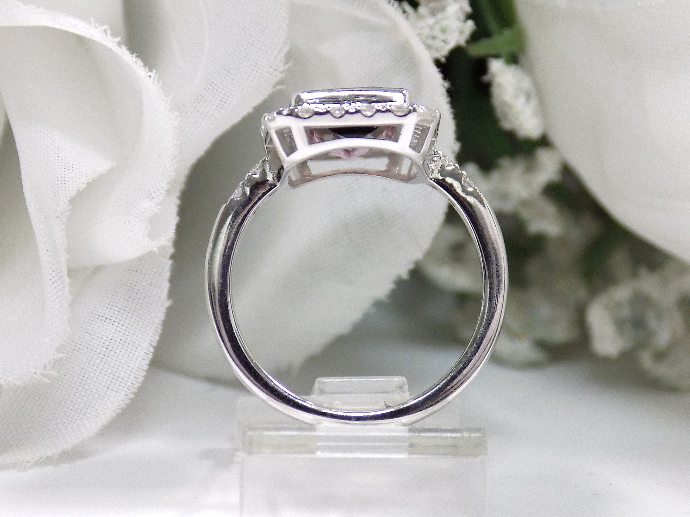 This is a women's ring with a natural 2.66ct bi-color tourmaline.  It's colors are a deep rich blueish green on one end that turns to a vivid pink on the other side.  The tourmaline is surrounded by .66ct in natural round accent diamonds in 14K white gold on a white surface with a white floral background from a side view.