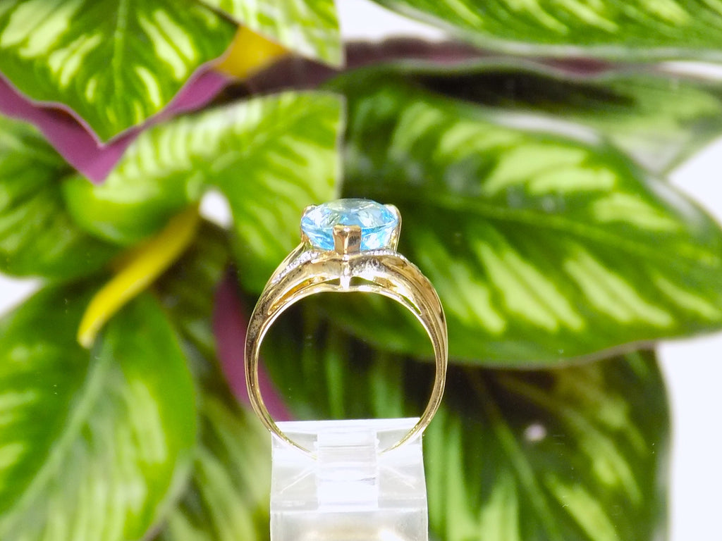 This is a side view of a genuine 7ct pear shape blue topaz ring in yellow gold from our estate collection in like new condition shown on mirrored finish with green leafy background.