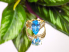 This is genuine 7ct pear shape blue topaz ring in yellow gold from our estate collection in like new condition shown on mirrored finish with green leafy background.