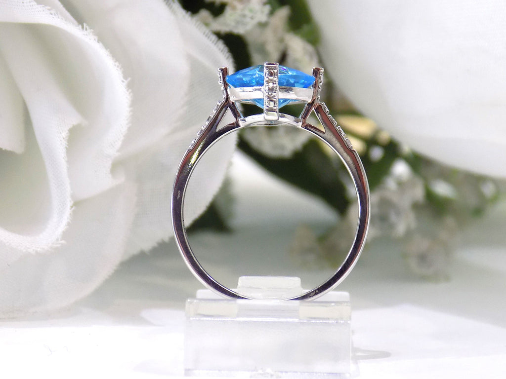 This is a diagonally set natural 2.50ct cushion cut blue topaz and .10ct diamond accents in 10K white gold side view with a white floral background.