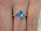 This is a diagonally set natural 2.50ct cushion cut blue topaz and .10ct diamond accents in 10K white gold on a womans finger.