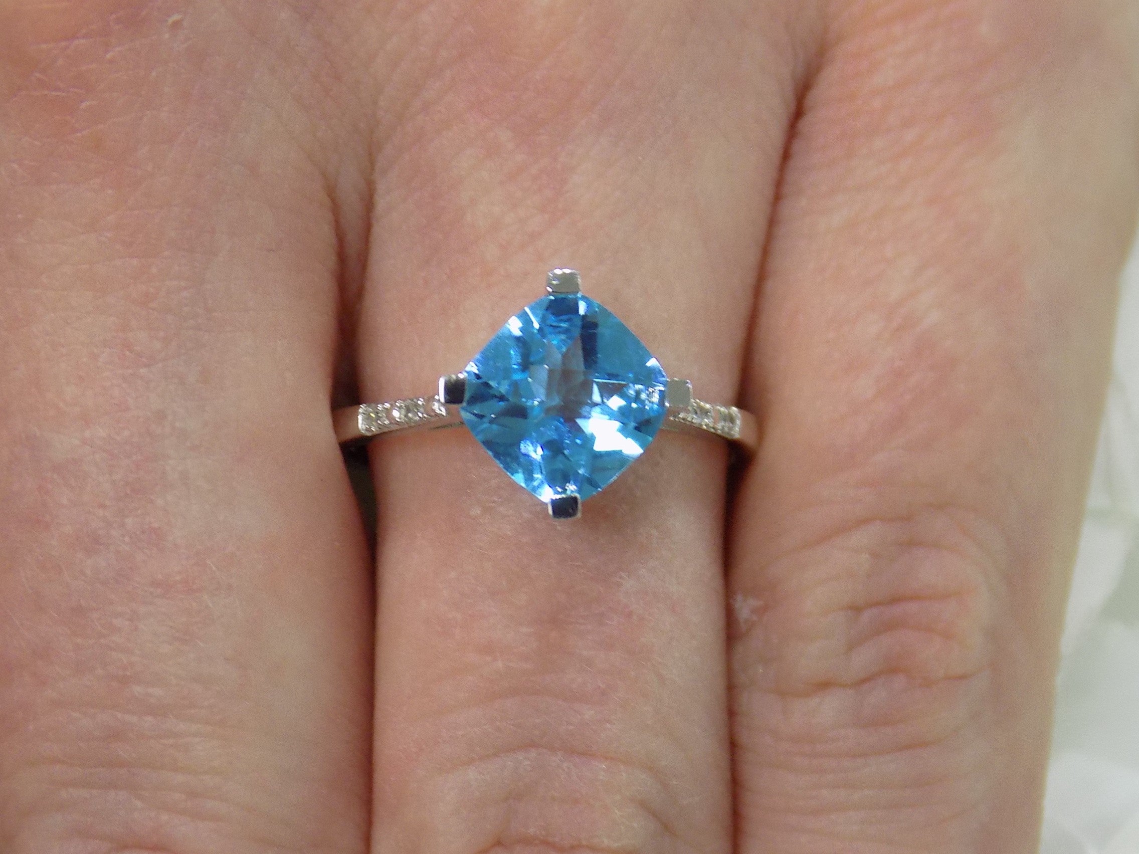This is a diagonally set natural 2.50ct cushion cut blue topaz and .10ct diamond accents in 10K white gold on a womans finger.