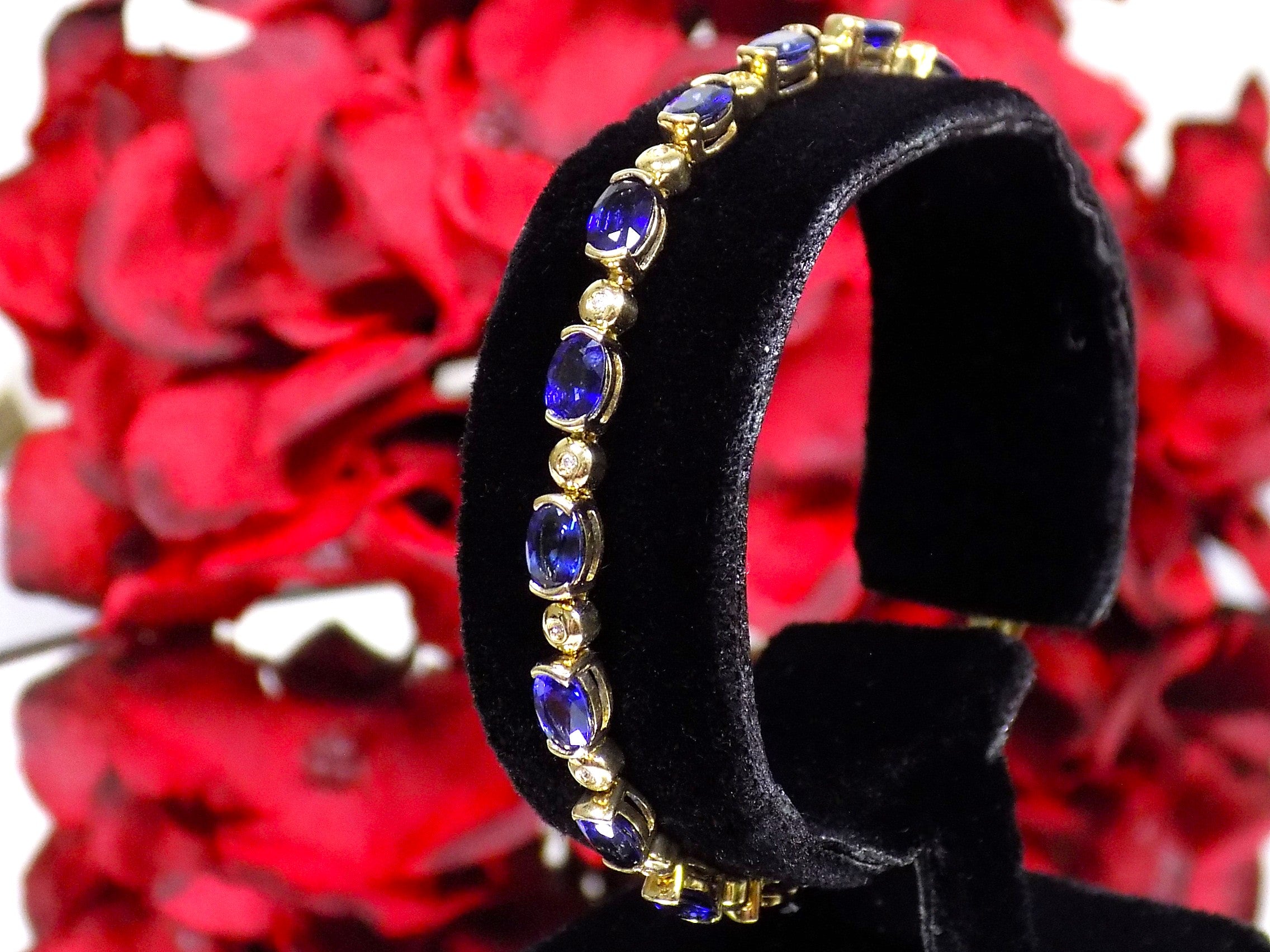 of natural oval shaped sapphires and round diamonds in 14K yellow gold on a black stand with a red floral background.