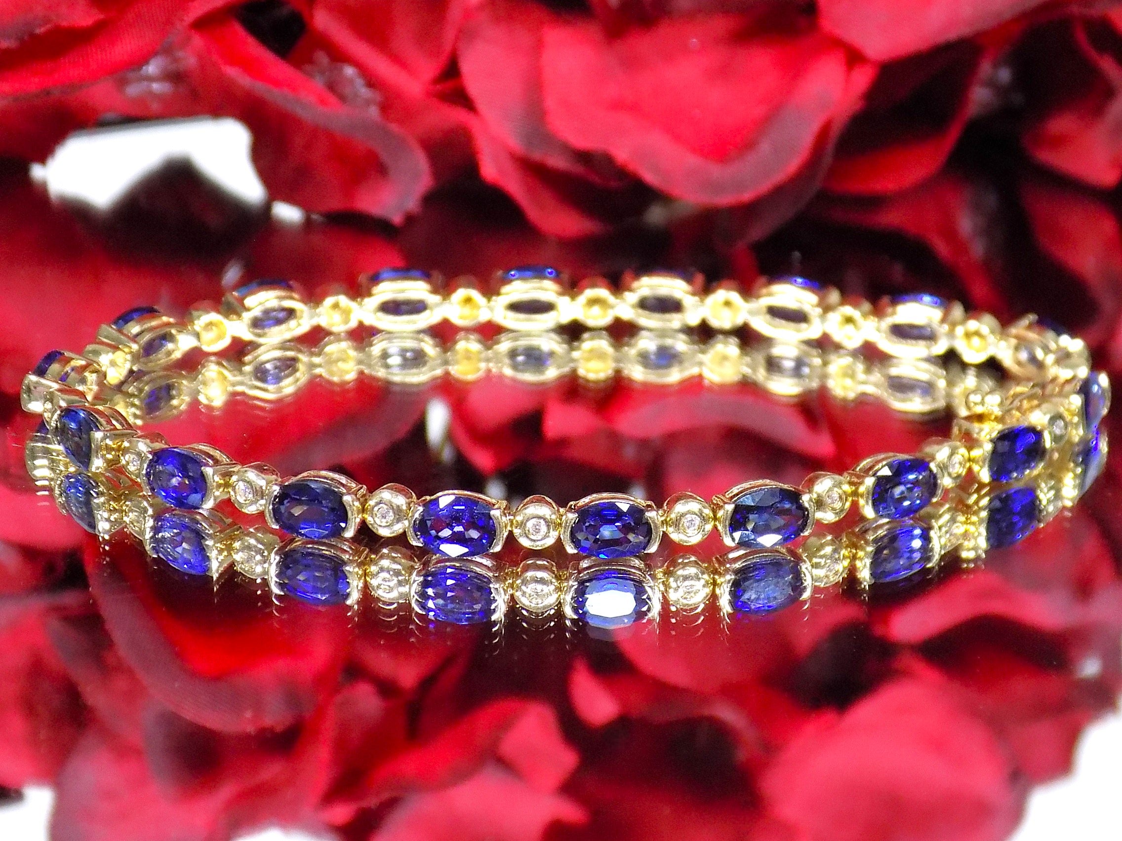 This is a women's bracelet with 10.00ct of natural oval shaped sapphires and round diamonds in 14K yellow gold on a mirrored surface with a red floral background.