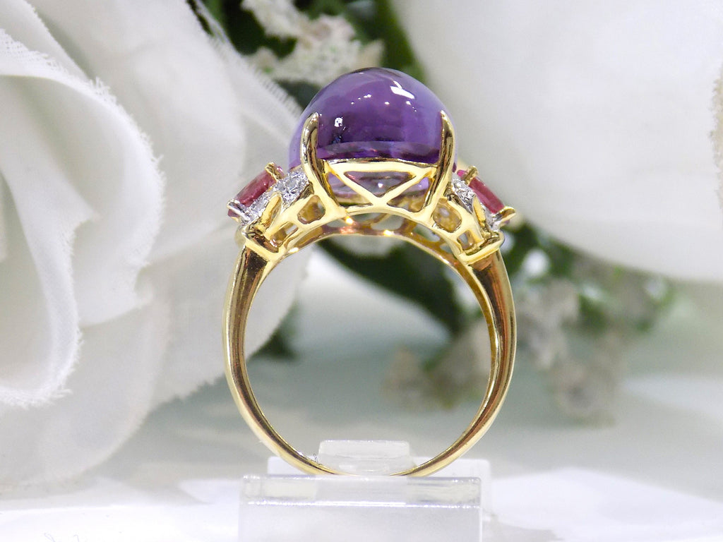 This is a women's natural 11.00ct cabochon shape amethyst with .20ct total weight natural pink tourmalines on either side plus .16ct total weight natural diamond accents on either side in 10K yellow gold with a white floral background from a side view.