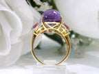 This is a women's natural 11.00ct cabochon shape amethyst with .20ct total weight natural pink tourmalines on either side plus .16ct total weight natural diamond accents on either side in 10K yellow gold with a white floral background from a side view.