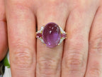 This is a women's natural 11.00ct cabochon shape amethyst with .20ct total weight natural pink tourmalines on either side plus .16ct total weight natural diamond accents on either side in 10K yellow gold on a women's finger.