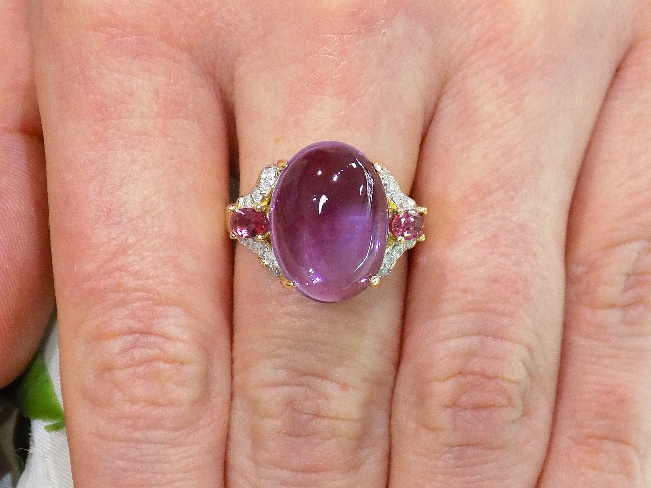 This is a women's natural 11.00ct cabochon shape amethyst with .20ct total weight natural pink tourmalines on either side plus .16ct total weight natural diamond accents on either side in 10K yellow gold on a women's finger.