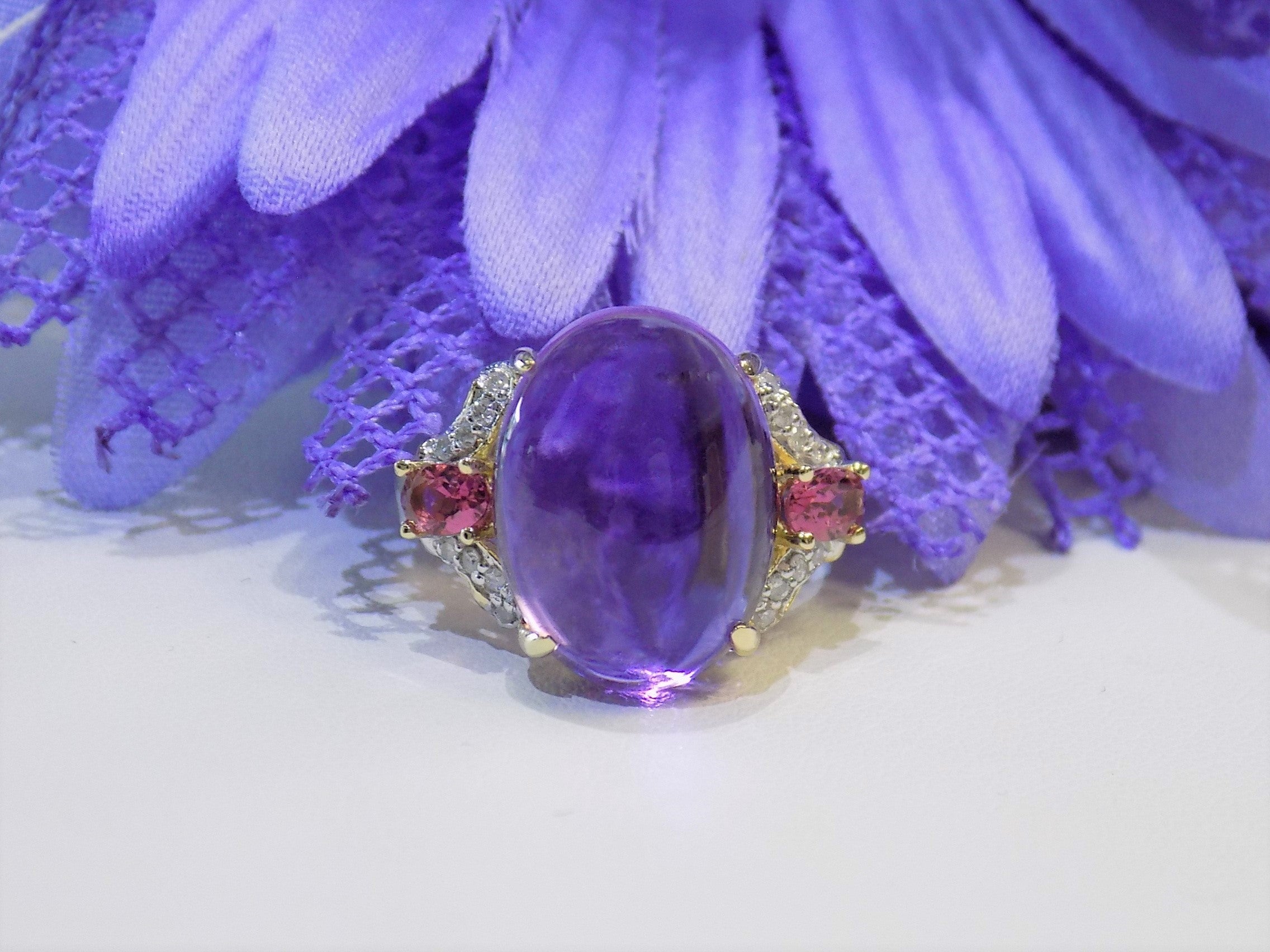 This is a women's natural 11.00ct cabochon shape amethyst with .20ct total weight natural pink tourmalines on either side plus .16ct total weight natural diamond accents on either side in 10K yellow gold with a purple floral background.