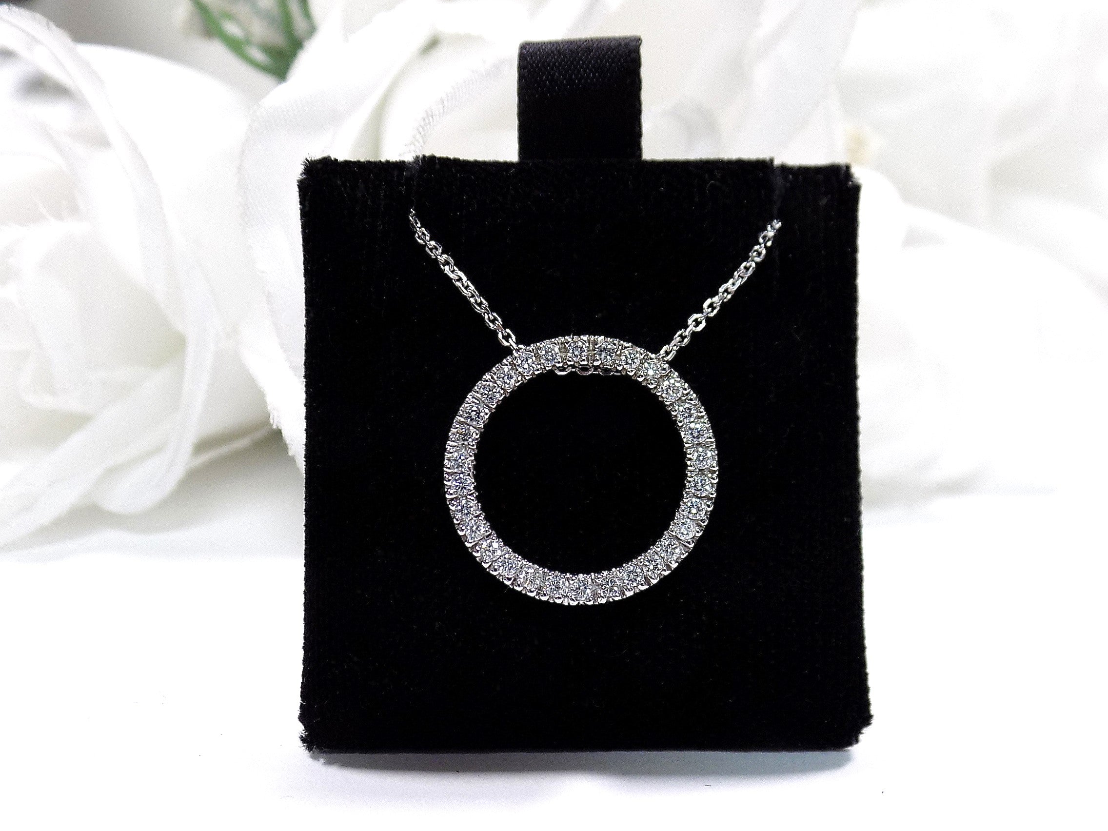 This is a circle .50ct natural diamond necklace with a cable style chain that can be adjusted in length of 14", 15", or 16" in 14K white gold displayed on a black pad with a white floral background.