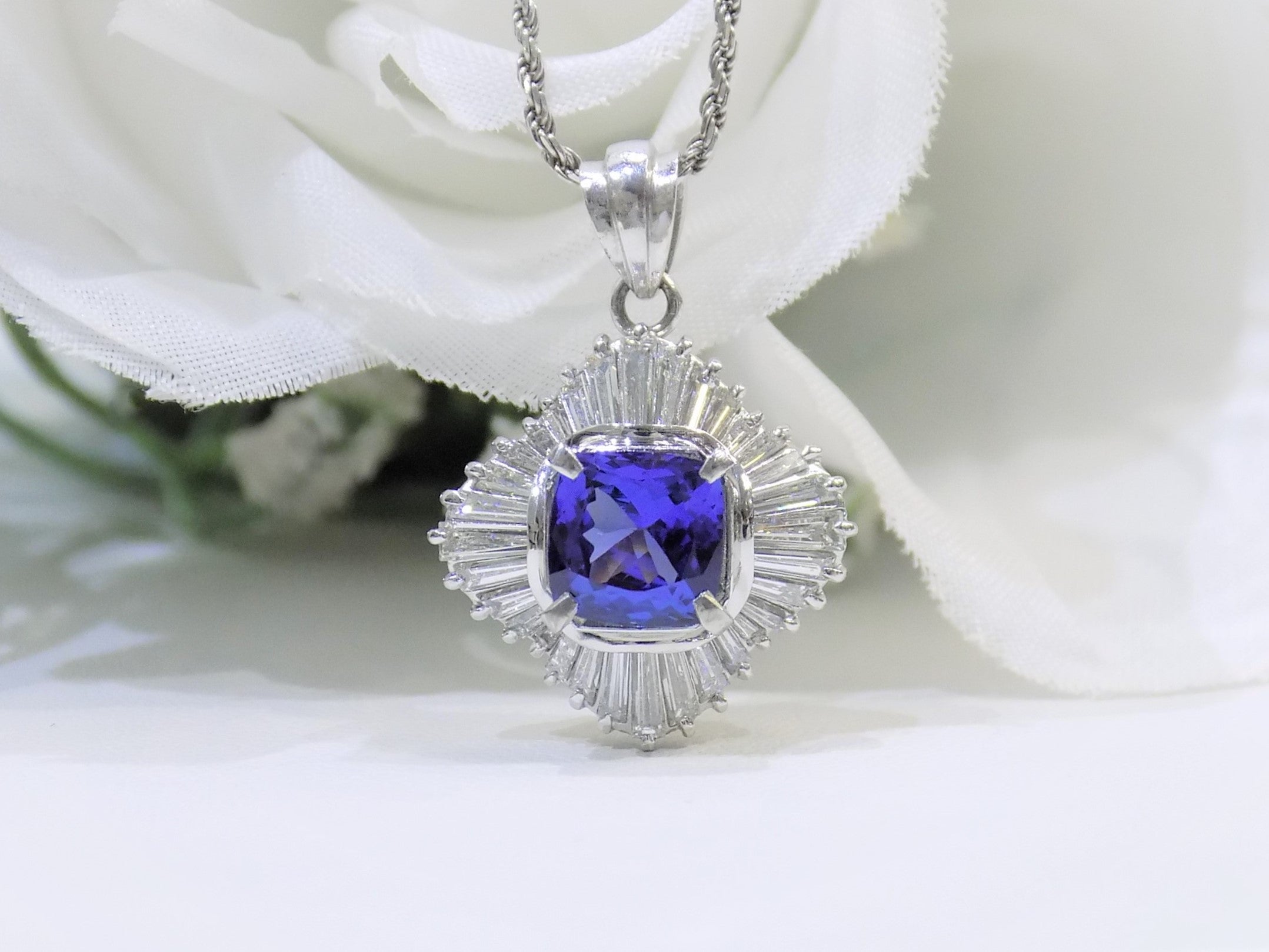 This is a women's ballerina/halo style pendant with a near perfect 2.04ct natural cushion cut tanzanite surrounded by a total weight of 1.06 natural baguette diamonds with a white floral background.