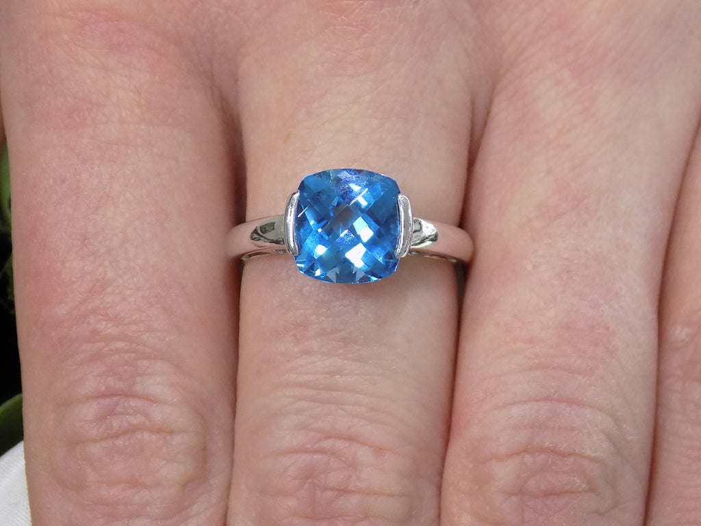 This is a tension set or channel set 4.00ct natural cushion cut blue topaz solitaire ring in 14K white gold on a ladies finger.