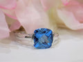 This is a tension set or channel set 4.00ct natural cushion cut blue topaz solitaire ring in 14K white gold on a white surface with a pink floral background.