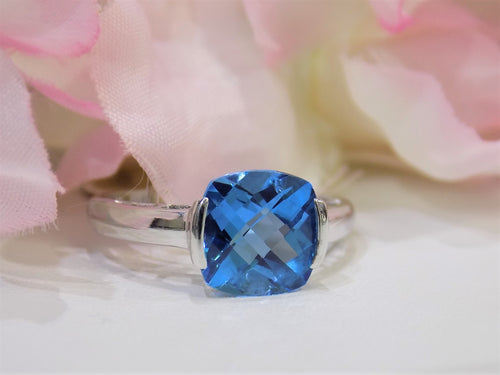 This is a tension set or channel set 4.00ct natural cushion cut blue topaz solitaire ring in 14K white gold on a white surface with a pink floral background.