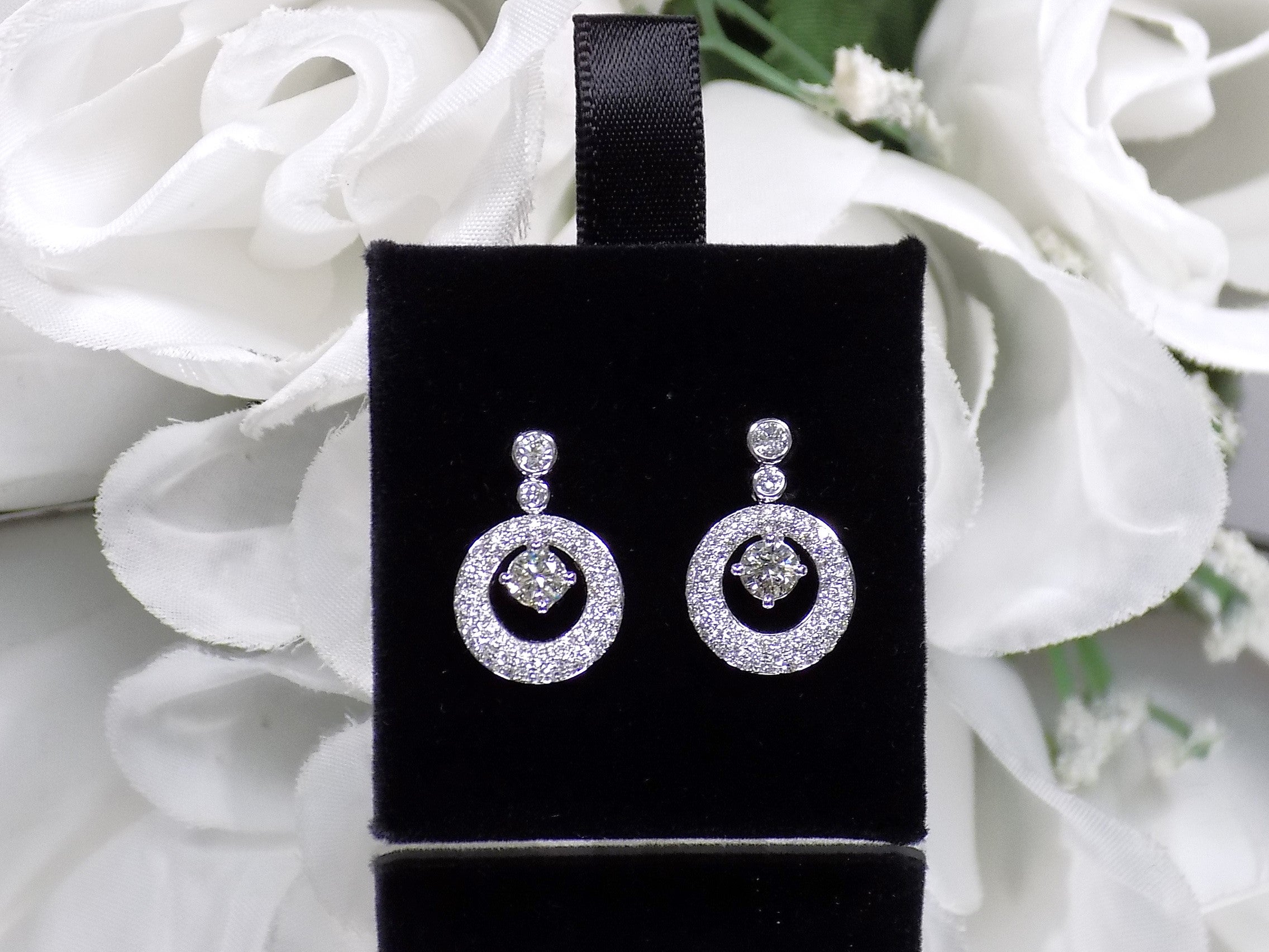 These are 1.34ct total weight natural round brilliant dancing diamond circle earrings in 14K white gold on a black pad with a floral background.