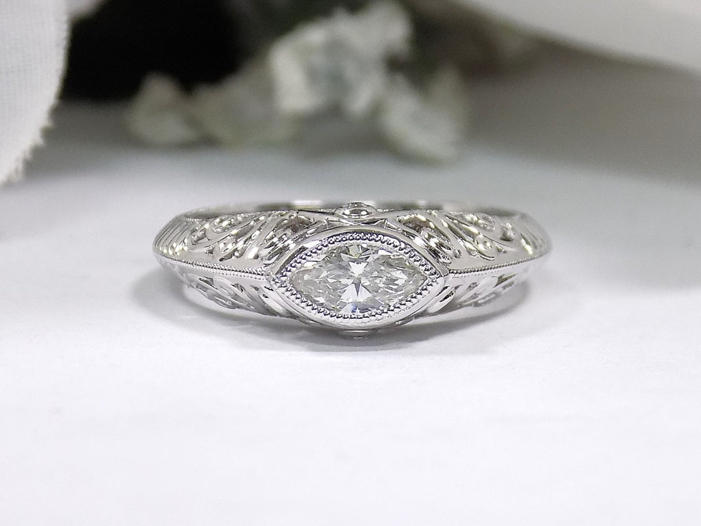 This is a bezel set marquise shape diamond set sideways in a white gold filigree ring with a white background.