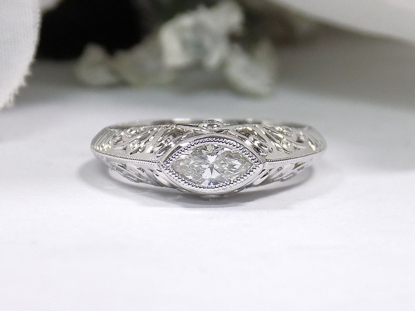 This is a bezel set marquise shape diamond set sideways in a white gold filigree ring with a white background.