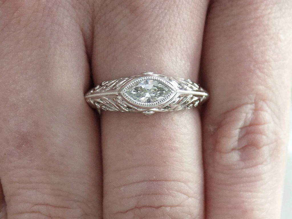 This is a bezel set marquise shape diamond set sideways in a white gold filigree mounting shown on a ladies' finger.