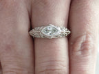 This is a bezel set marquise shape diamond set sideways in a white gold filigree mounting shown on a ladies' finger.
