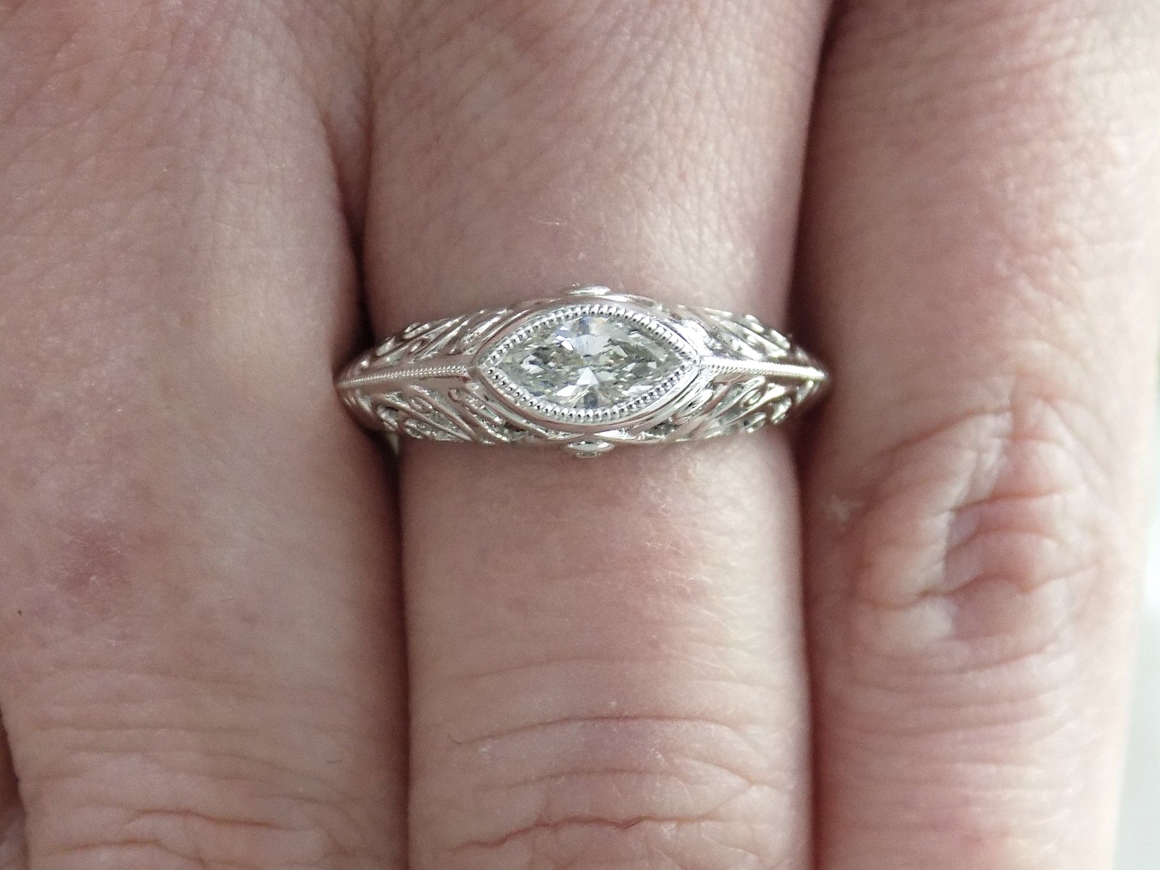 This is a bezel set marquise shape diamond set sideways in a white gold filigree mounting shown on a ladies' finger.