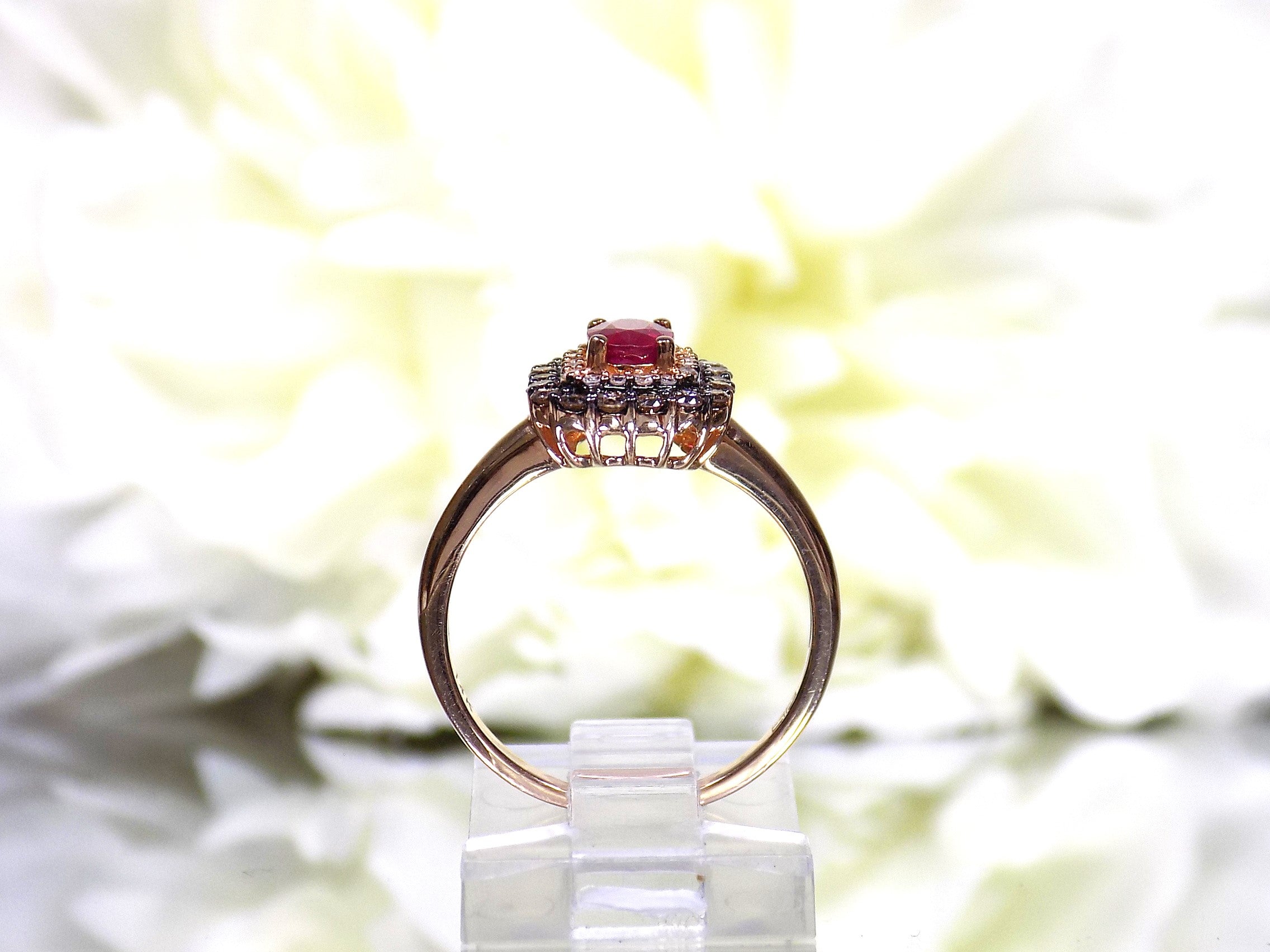 This is a women's effy brand ring with a .57ct natural oval shape ruby with a halo of natural white diamonds and a halo of chocolate diamonds on the outside for a total wight of .43ct in diamonds set in 14K rose gold with a white floral background in a side view.