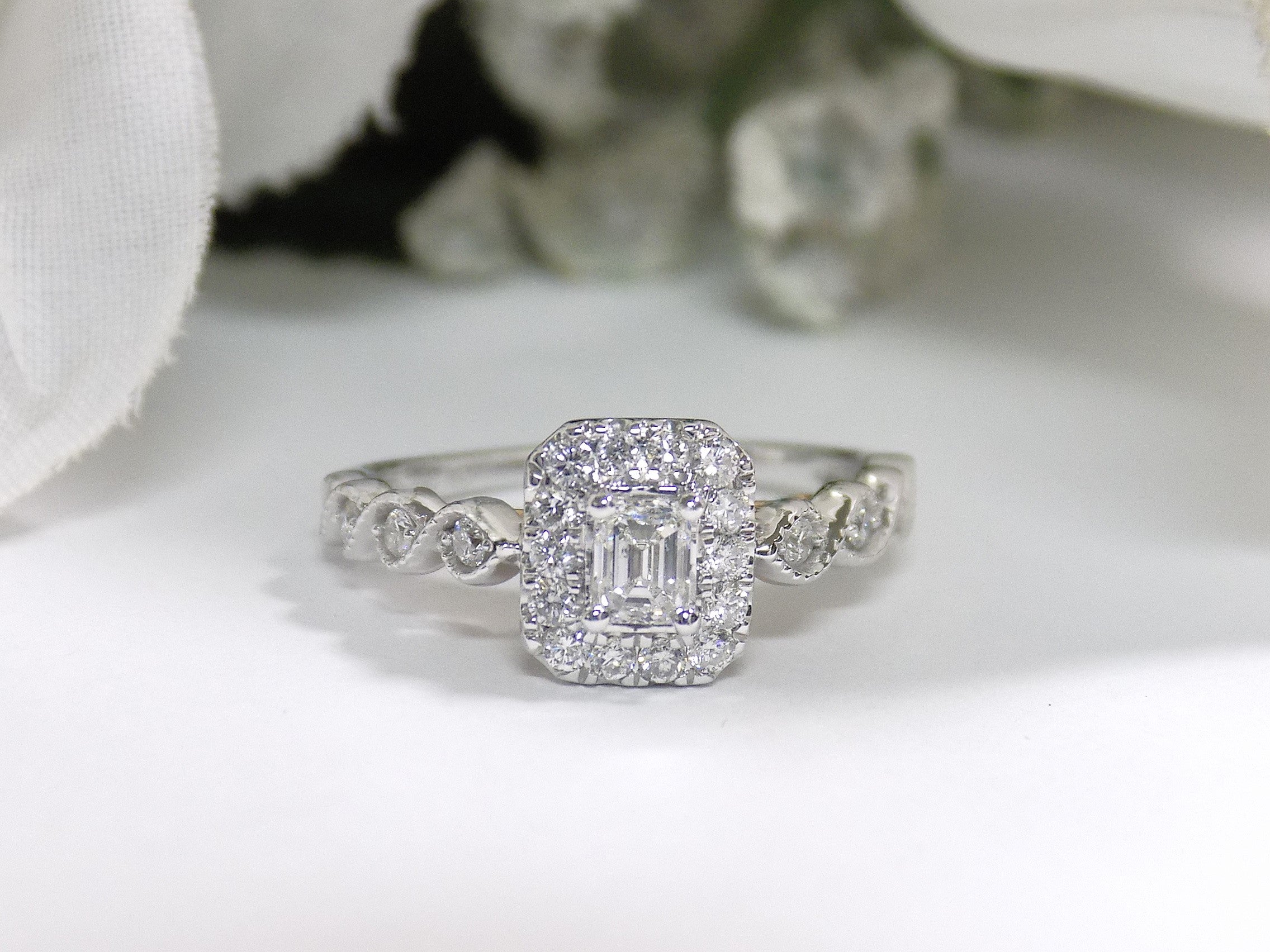 This is a .75ct natural emerald cut halo style ring with infinity style band in white gold with rose gold accents on the sides with a white background.