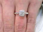 This is a .75ct natural emerald cut halo style ring with infinity style band in white gold shown on a woman's finger.
