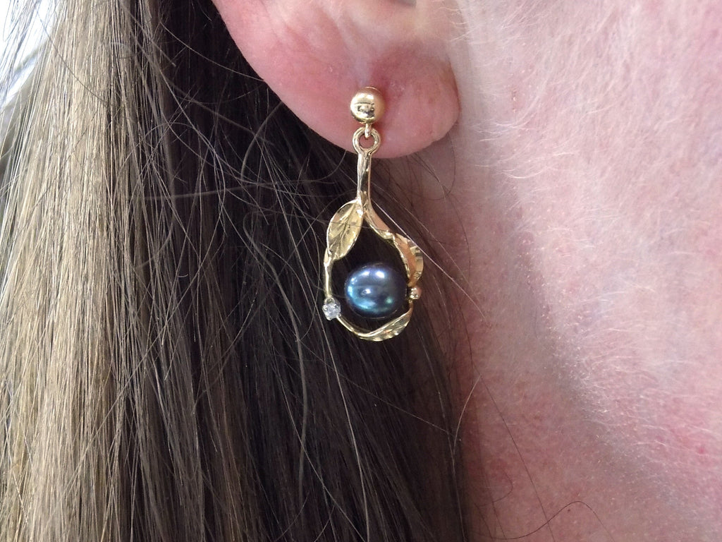 This is a pair of genuine black pearl earrings in yellow gold shown in a woman's ear.