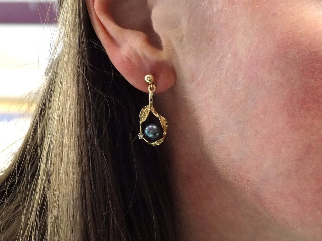 This is a pair of genuine black pearl earrings in yellow gold shown in a woman's ear.