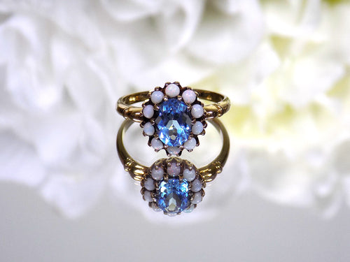 This a ring with an oval blue topaz in the center and surrounded by natural opals in yellow gold from our estate collection with a mirrored flowery background.