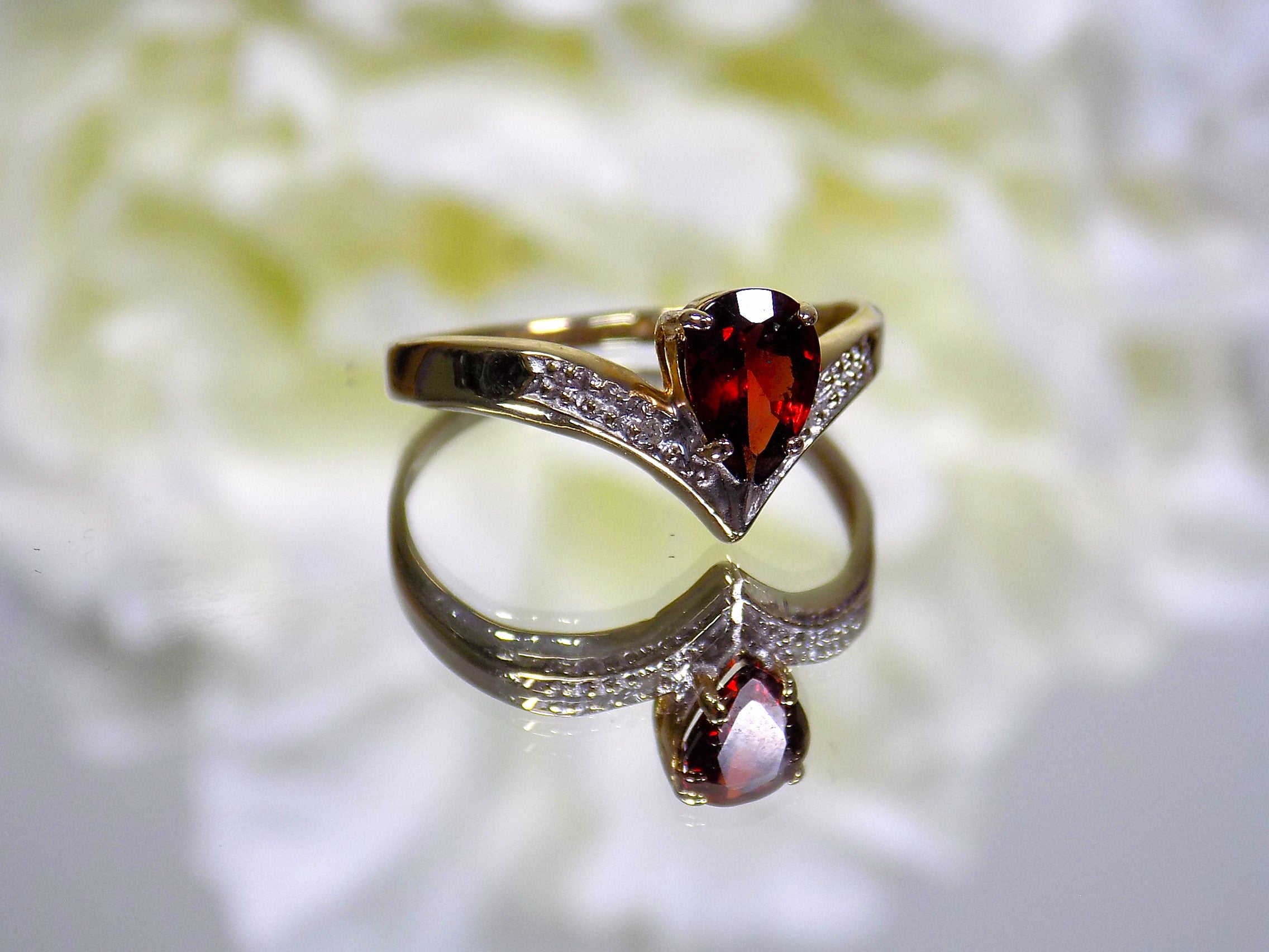 This is a pear shape garnet ring with diamond accents in yellow gold from our estate collection with flowery mirrored background.