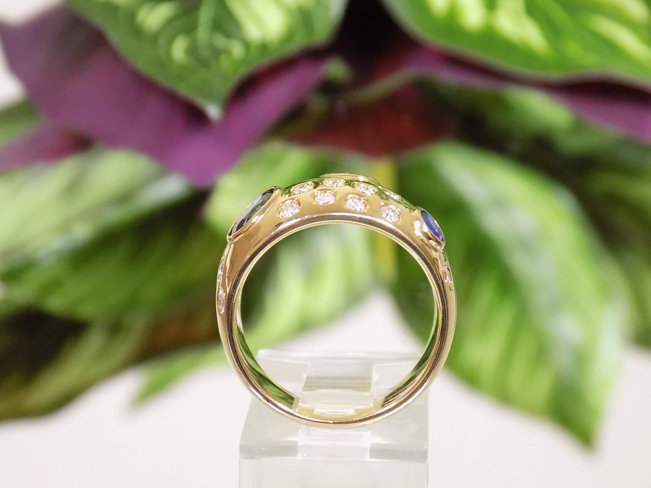 This is a women's estate ring with 2.50ct total weight ruby, sapphire, and diamonds set in 18K yellow gold with a green leafy background from a side view.
